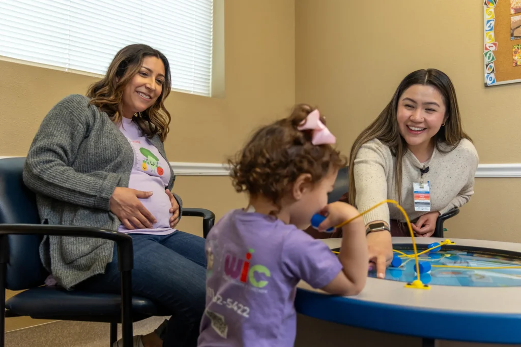 A pregnant woman sits smiling beside a young girl playing with a bead maze toy, while another woman with a name badge interacts with the child in a brightly lit, welcoming room.